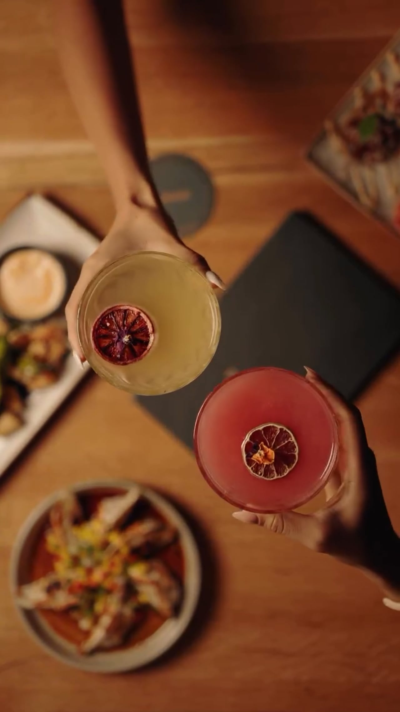 Two cocktails with dried citrus wheels raised over a table of shared dishes